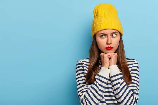 Foto de estudio de pensativa modelo de mujer joven mira a un lado con expresión seria, vestida con suéter a rayas, elegante sombrero amarillo, aislado sobre fondo azul, espacio en blanco para promo