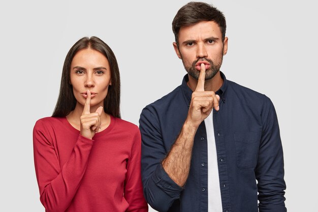 Foto de estudio de secreto joven mujer caucásica y el hombre mantienen los dedos índices en los labios, parados uno al lado del otro