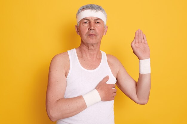 Foto de estudio de serio hombre maduro concentrado posando contra la pared amarilla, vistiendo camiseta blanca sin mangas y banda para la cabeza