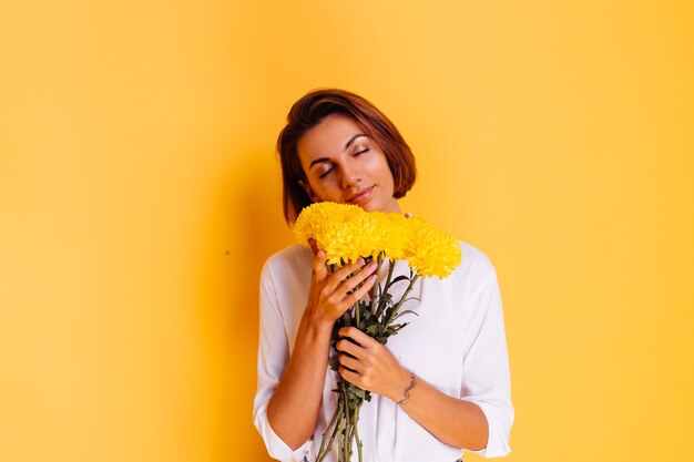 Foto de estudio sobre fondo amarillo Feliz mujer caucásica de pelo corto con ropa casual camisa blanca y pantalones de mezclilla con ramo de asters amarillos