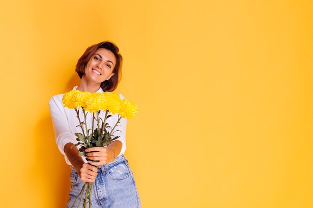 Foto de estudio sobre fondo amarillo Feliz mujer caucásica de pelo corto con ropa casual camisa blanca y pantalones de mezclilla con ramo de asters amarillos