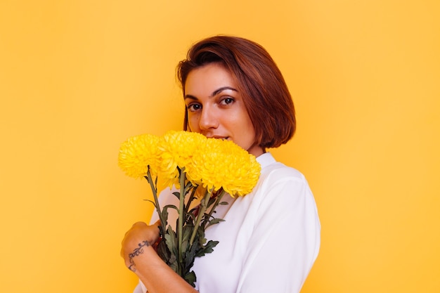 Foto de estudio sobre fondo amarillo Feliz mujer caucásica de pelo corto con ropa casual camisa blanca y pantalones de mezclilla con ramo de asters amarillos