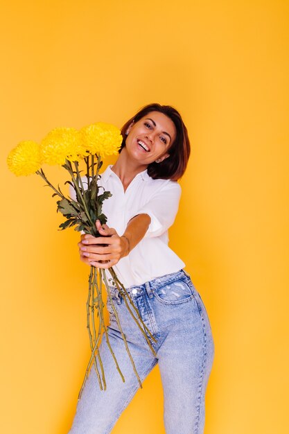 Foto de estudio sobre fondo amarillo Feliz mujer caucásica de pelo corto con ropa casual camisa blanca y pantalones de mezclilla con ramo de asters amarillos