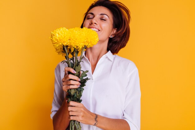 Foto de estudio sobre fondo amarillo Feliz mujer caucásica de pelo corto con ropa casual camisa blanca y pantalones de mezclilla con ramo de asters amarillos