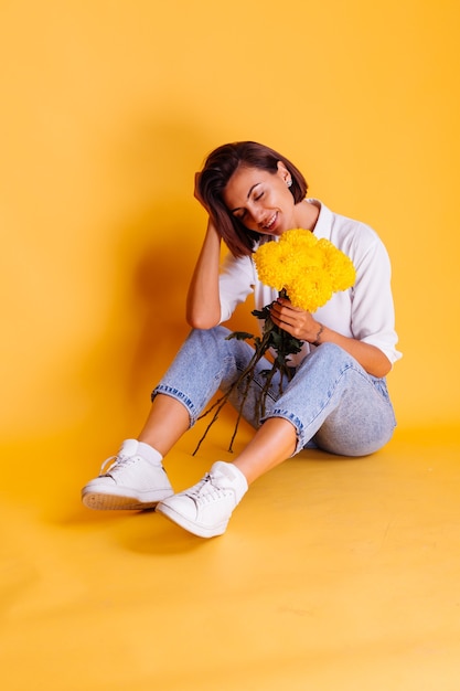 Foto gratuita foto de estudio sobre fondo amarillo feliz mujer caucásica de pelo corto con ropa casual camisa blanca y pantalones de mezclilla con ramo de asters amarillos