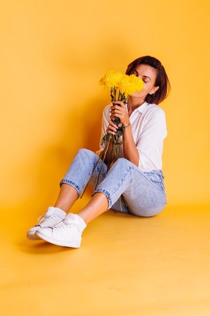 Foto de estudio sobre fondo amarillo Feliz mujer caucásica de pelo corto con ropa casual camisa blanca y pantalones de mezclilla con ramo de asters amarillos