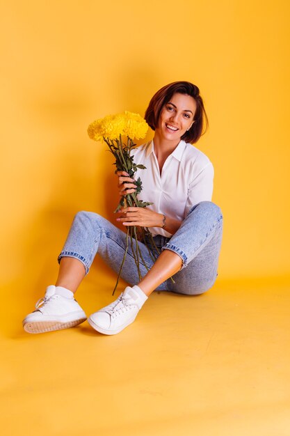 Foto de estudio sobre fondo amarillo Feliz mujer caucásica de pelo corto con ropa casual camisa blanca y pantalones de mezclilla con ramo de asters amarillos