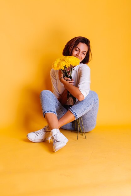 Foto de estudio sobre fondo amarillo Feliz mujer caucásica de pelo corto con ropa casual camisa blanca y pantalones de mezclilla con ramo de asters amarillos