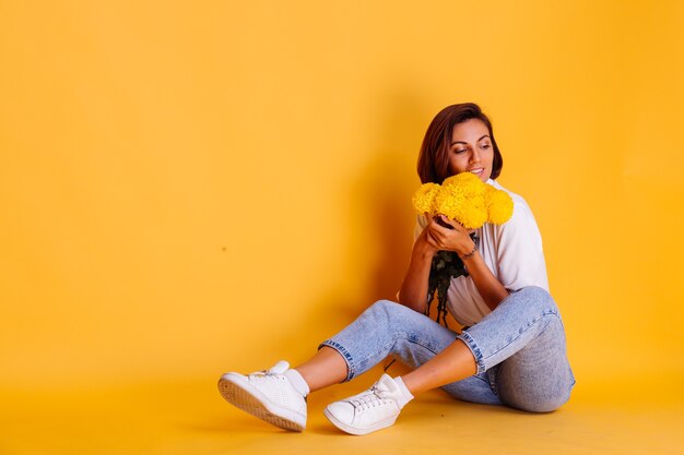 Foto de estudio sobre fondo amarillo Feliz mujer caucásica de pelo corto con ropa casual camisa blanca y pantalones de mezclilla con ramo de asters amarillos