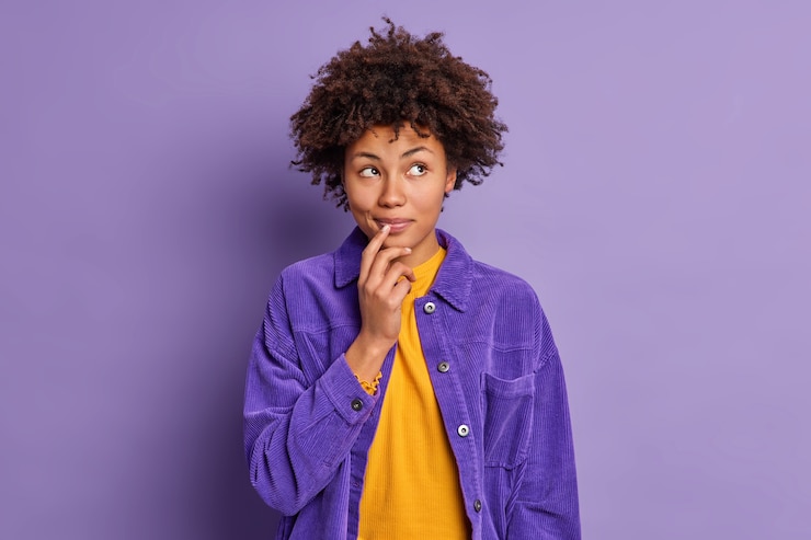 Foto gratuito foto de estudio de soñadora joven de piel oscura mantiene las manos cerca de la boca mira pensativamente a un lado vestida con elegante chaqueta violeta