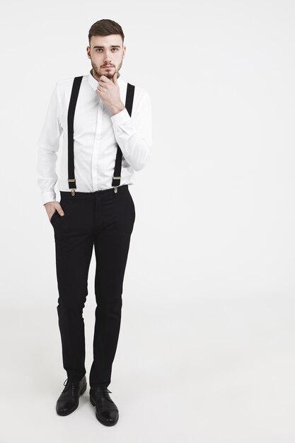 Foto de estudio vertical de longitud completa de un elegante y guapo modelo masculino barbudo joven con pantalón negro, zapatos y camisa blanca con tirantes tocando su barba, posando para el catálogo de ropa masculina