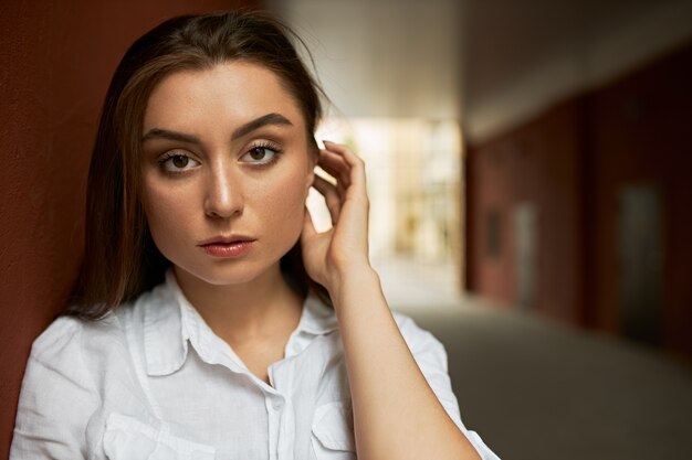 Foto de una exitosa joven empresaria atractiva con camisa blanca poniendo el pelo detrás de la oreja, mirando a la cámara con expresión facial seria y segura