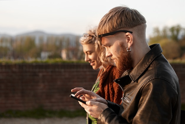 Foto exterior de moda joven pelirrojo con pendiente y chaqueta de motorista con teléfono móvil mientras camina junto con una hermosa chica rubia. Concepto de primera cita, romance y tecnología.