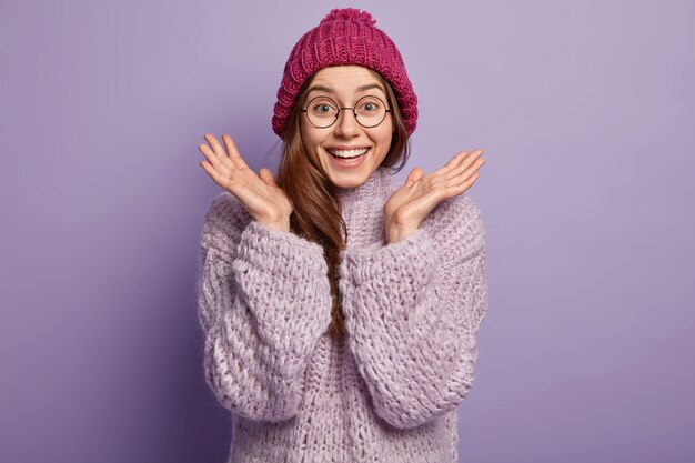 Foto de felices gestos de mujer europea emotiva con ambas manos, abrocha las palmas, lleva gafas redondas, vestida con gorro de invierno, jersey de punto, aislado sobre una pared púrpura Concepto de buenas emociones