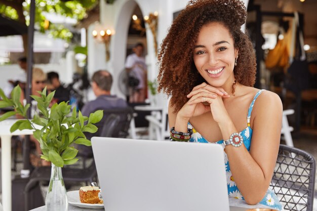 Foto de feliz adorable mujer africana rizada sentada frente a la computadora portátil abierta en la cafetería en la acera, satisfecha de hacer una buena presentación