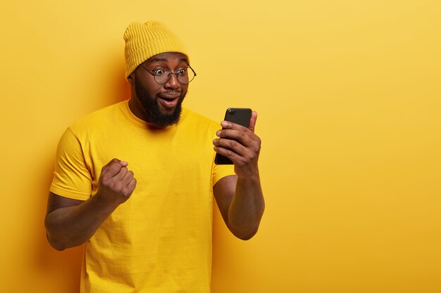 Foto de feliz chico de piel oscura celebra la victoria de su equipo favorito, lee los resultados del juego en Internet, se ve muy feliz en la pantalla del teléfono inteligente