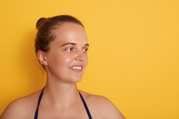 Foto de feliz joven mujer de raza blanca con tierna sonrisa, mira a un lado con expresión encantadora, viste traje de baño, tiene belleza natural, aislado sobre la pared amarilla.