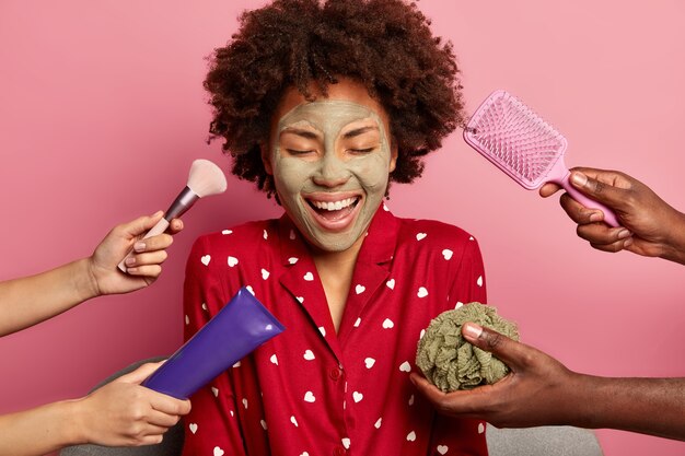 Foto de feliz joven de piel oscura aplica mascarilla de arcilla purificante, viste pijama rojo, tiene los ojos cerrados, recibe tratamientos cosméticos