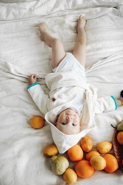 Foto genérica de cuerpo entero de un lindo joven en bata de baño blanca tendido descalzo sobre una cama blanca con frutas esparcidas y sonriendo a la cámara. Limones, peras y naranjas esparcidos sobre la cama en la cabeza del niño.