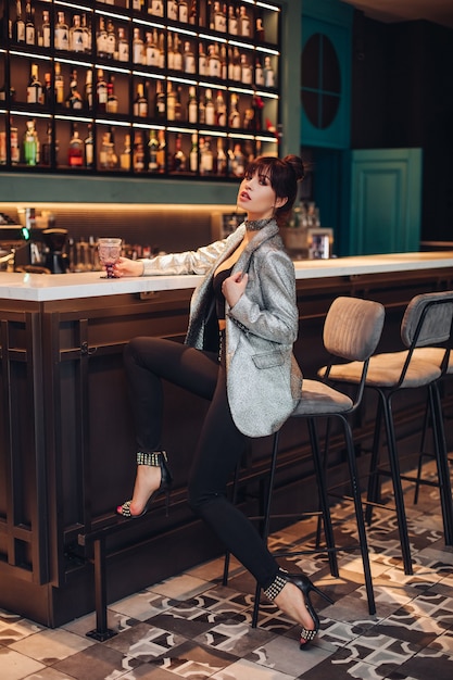 Foto genérica de cuerpo entero de una modelo morena lujosa extremadamente moderna en top corto, chaqueta plateada brillante, pantalón negro y tacones altos. Modelo en traje de moda sentado en taburete de bar en club o bar.