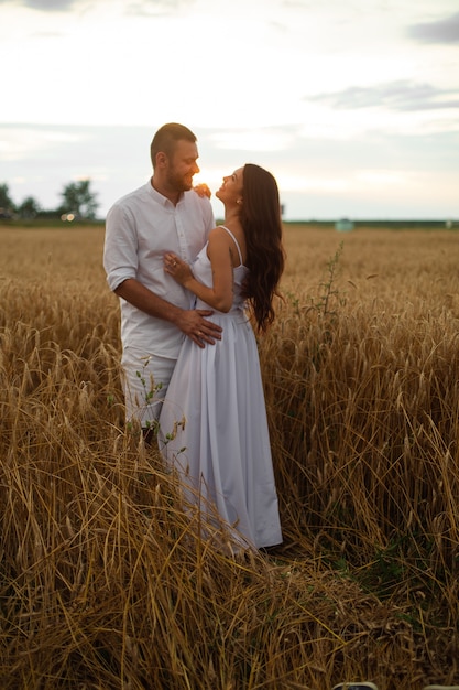 Foto genérica de longitud completa de una pareja romántica en ropa blanca abrazándose en el campo de trigo al atardecer.