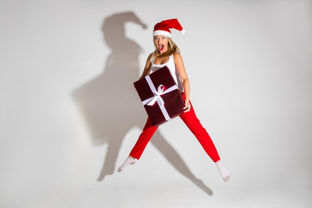 Foto genérica de saltar alegre mujer rubia en rojo sombrero de Papá Noel y pantalones rojos con un regalo de Navidad envuelto con la boca abierta.