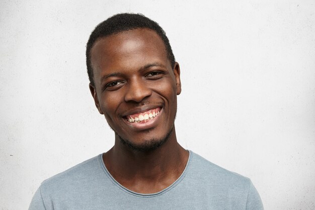 Foto de guapo joven afroamericano mirando con una amplia sonrisa amistosa, disfrutando de buenos días y tiempo libre en el interior. Hombre negro sentirse feliz y despreocupado mientras se relaja en casa