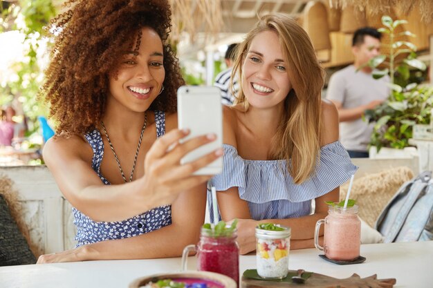 Foto de hembras de raza mixta alegres que tienen amistad interracial, posan en la cámara del teléfono celular moderno, hacen selfie mientras descansan en el acogedor bar de la terraza, disfrutan de bebidas frescas. Gente, etnia y ocio