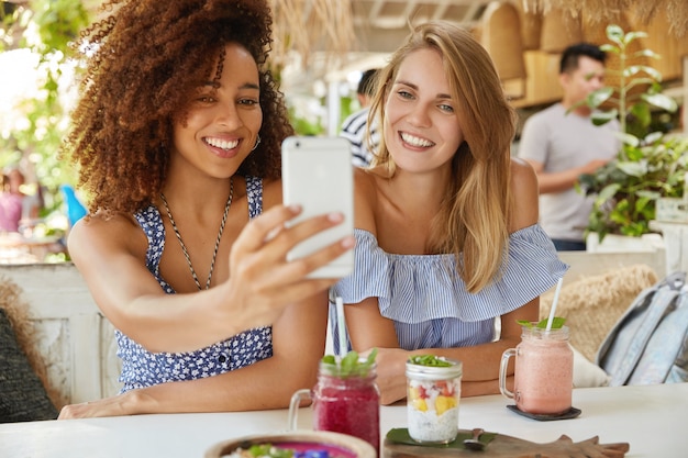 Foto de hembras de raza mixta alegres que tienen amistad interracial, posan en la cámara del teléfono celular moderno, hacen selfie mientras descansan en el acogedor bar de la terraza, disfrutan de bebidas frescas. Gente, etnia y ocio