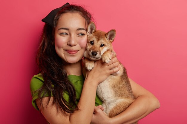 Foto de una hermosa chica coreana enamorada de su perro shiba inu, abraza a la mascota con una sonrisa, tiene el pelo oscuro, viste una camiseta verde, posa con un animal sobre un fondo rosa.