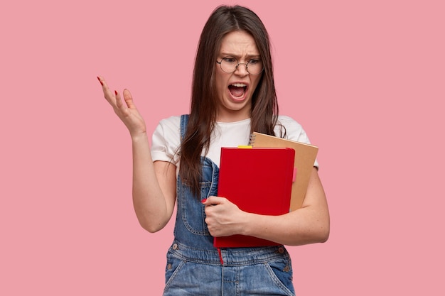 Foto de una hermosa colegiala indignada levanta la mano, exclama con irritación, mira el bloc de notas y el libro de texto, molesta por estudiar