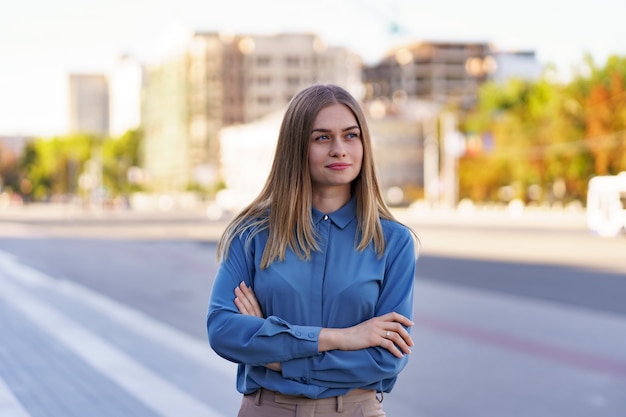 Foto de hermosa joven empresaria vistiendo camisa azul chifon mientras está de pie en la calle con los brazos cruzados.