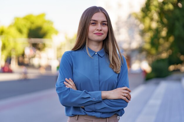 Foto de hermosa joven empresaria vistiendo camisa azul chifon mientras está de pie en la calle con los brazos cruzados.