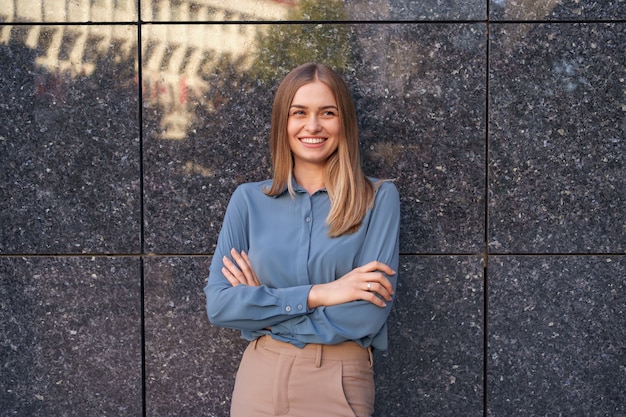 Foto de hermosa joven empresaria vistiendo camisa de gasa azul mientras está de pie con los brazos cruzados sobre la pared de mármol gris