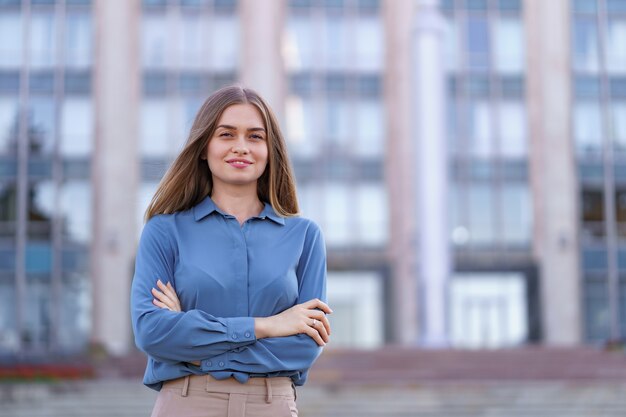 Foto de hermosa joven empresaria vistiendo camisa de gasa azul mientras está de pie en el edificio en la calle con los brazos cruzados.