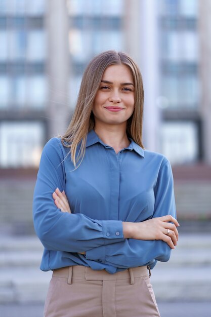 Foto de hermosa joven empresaria vistiendo camisa de gasa azul mientras está de pie en el edificio en la calle con los brazos cruzados.