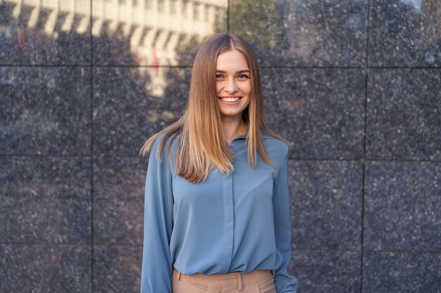 Foto de hermosa joven empresaria vistiendo camisa de gasa azul mientras está de pie y posando en la pared de mármol gris