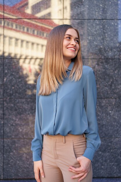 Foto de hermosa joven empresaria vistiendo camisa de gasa azul mientras está de pie y posando en la pared de mármol gris