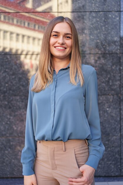 Foto de hermosa joven empresaria vistiendo camisa de gasa azul mientras está de pie y posando en la pared de mármol gris