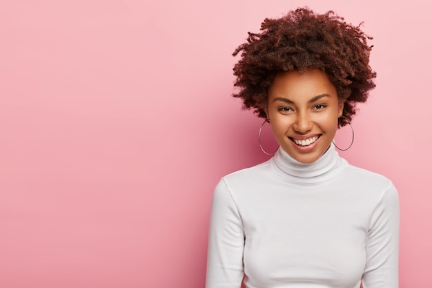 Foto de una hermosa jovencita con cabello afro rizado, sonríe suavemente, usa aretes y un jersey blanco, está satisfecha con conseguir un nuevo puesto de trabajo, tiene una conversación agradable con su colega, se para sobre una pared rosa