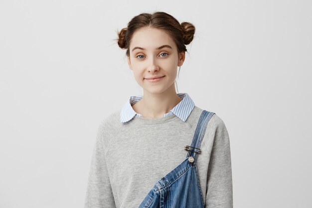 Foto de hermosa mujer de 20 años con aspecto europeo de pie sobre la pared blanca sonriendo sinceramente. Actriz femenina en overol de mezclilla que asiste al casting para una nueva película. Concepto de felicidad