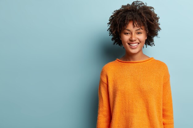 Foto de una hermosa mujer afroamericana positiva con cabello rizado, viste un jersey naranja, estando de buen humor se encuentra sobre una pared azul con espacio libre para su contenido promocional. Buen concepto de emociones