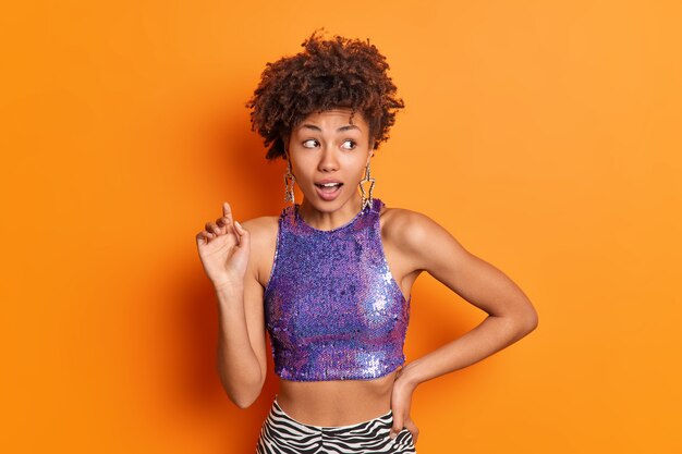 Foto de hermosa mujer afroamericana sorprendida mantiene la mano en la cintura sorprendentemente a un lado