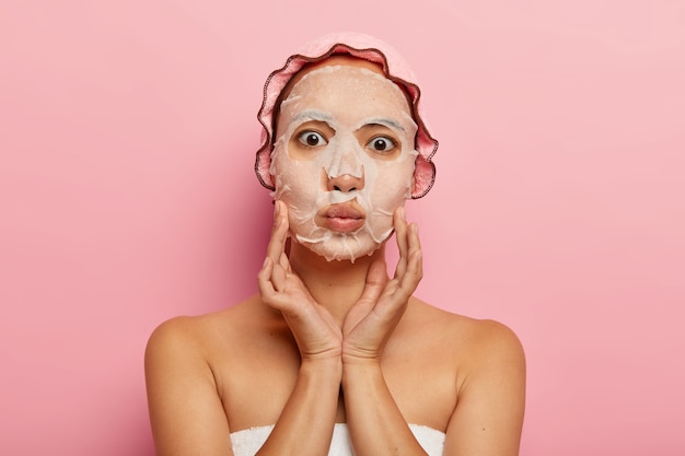 La foto de una hermosa mujer asiática tiene una máscara de papel humectante nutritiva en la cara, toca las mejillas suavemente, mantiene los labios doblados, usa un gorro de ducha rosa, está sola. Concepto de tratamientos de belleza y cuidado de la piel.