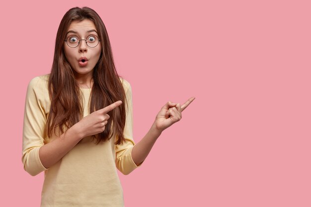 Foto de hermosa mujer asombrada con expresión de asombro apunta a un espacio en blanco, demuestra algo terrible, vestida con ropa casual