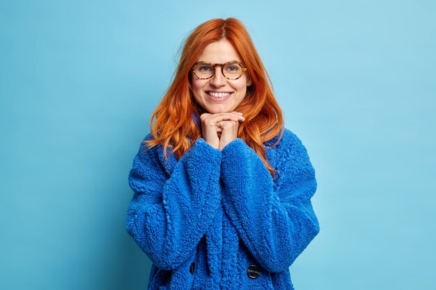 Foto de hermosa mujer europea pelirroja sonríe con alegría y mantiene las manos debajo de la barbilla lleva gafas ópticas y abrigo de piel caliente.
