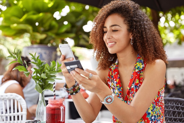 Foto de hermosa mujer joven alegre con peinado afro, número de tipos de tarjeta de crédito en el teléfono inteligente, realiza compras en línea o verifica la cuenta bancaria