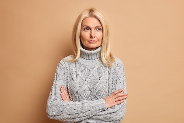 Foto de hermosa mujer de mediana edad con cabello rubio mantiene los brazos cruzados, piensa en algo que contempla sobre planes futuros y usa un suéter cálido