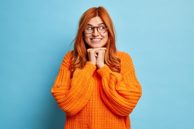 Foto de hermosa mujer pelirroja mantiene las manos debajo de la barbilla y sonríe sinceramente mira a un lado con expresión soñadora usa anteojos tejidos de color naranja.