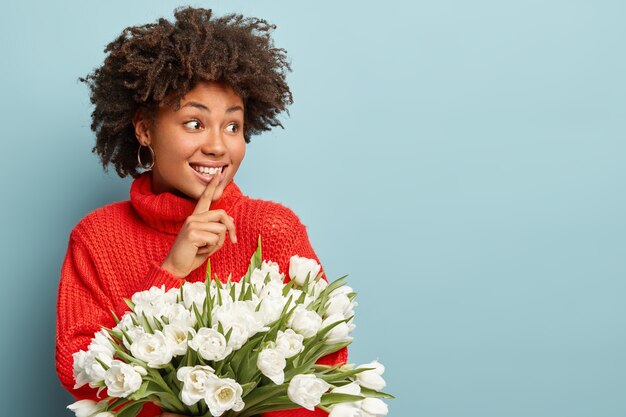 Foto de hermosa mujer rizada mira alegremente a un lado, hace gesto de silencio, vestida con un suéter rojo de invierno, dice el secreto quién dio tulipanes blancos, chismes con un amigo, aislado en azul Señora sostiene flores